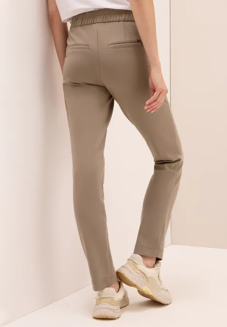 Person in beiger Hose, weißen Sneakers und weißem Shirt lehnt an der Wand.